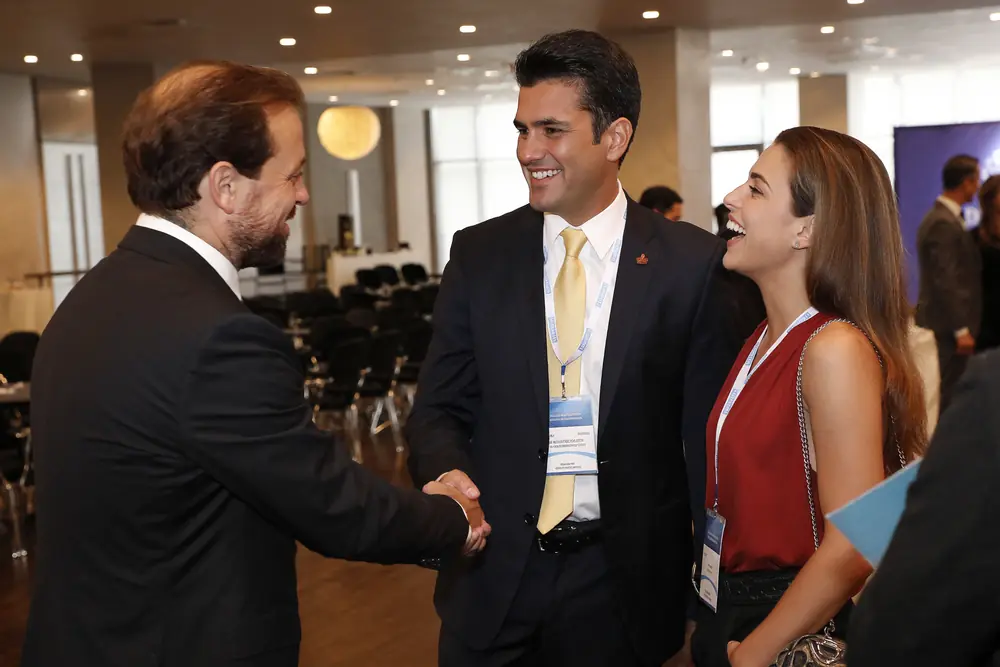 Two businessmen shaking hands at an event, next to a smiling businesswoman